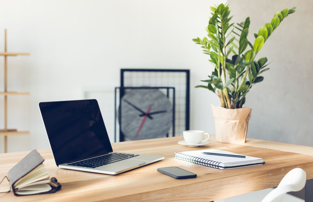 A clean and organized desk showing a MacBook on a brown desk with a plant, a notebook, a phone, and a cup of coffee.